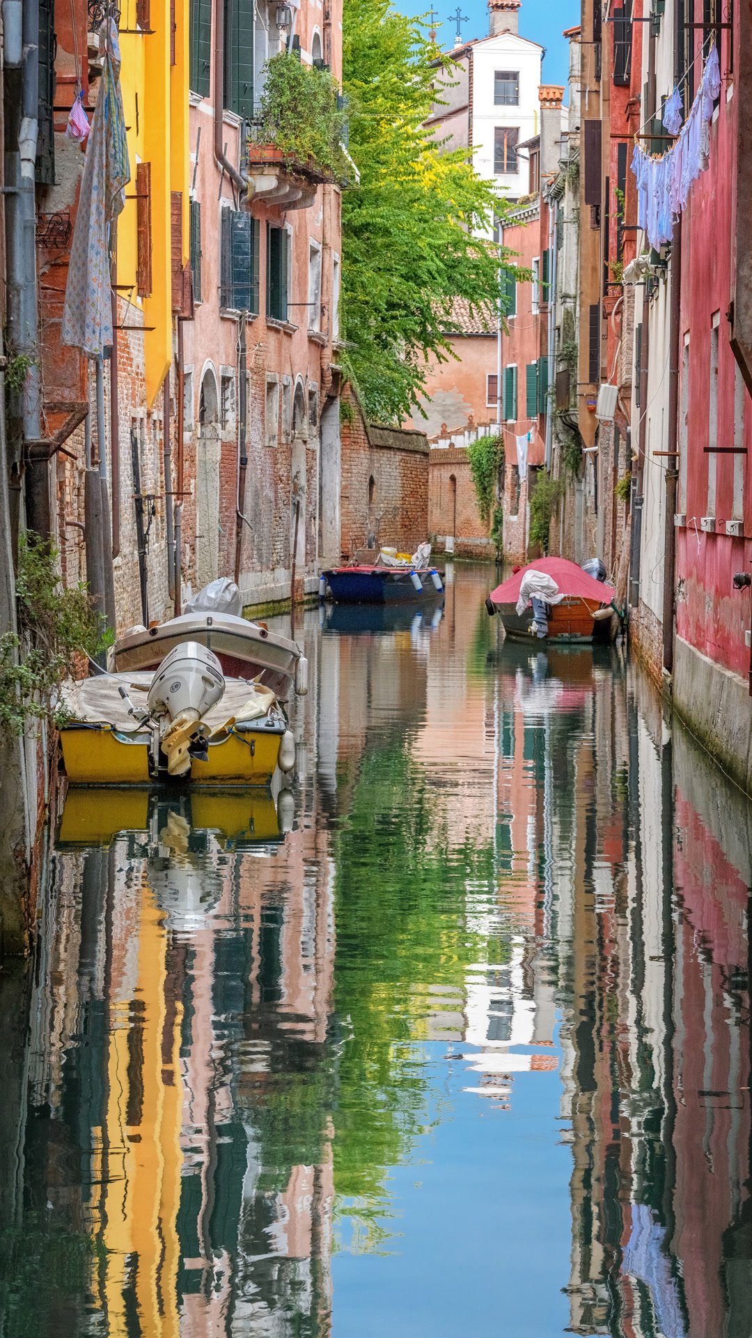 A narrow canal with moored boats and coloured houses reflected in the water.